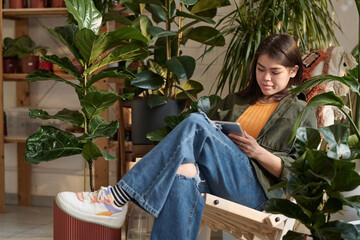 Female florist worker communicating with clients via tablet sitting in wooden chair amid houseplants