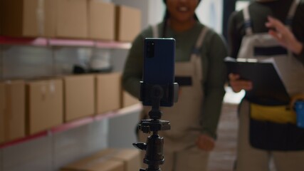 Warehouse logistics coordinator and colleague using smartphone placed on tripod to make training video for interns. Employees film themselves in fulfillment center showing trainees how to seal boxes