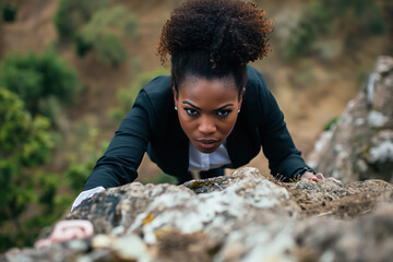 Gender and racial equality of success - black businesswoman struggle to climb up a mountain - front face view of pulling herself up - obrazy, fototapety, plakaty