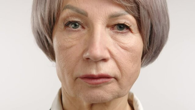 Intense high-definition close-up view of a senior woman's face with short grey hair, piercing blue eyes, and a thoughtful expression, showcasing detailed textures and a serene ambiance. Camera 8K RAW.