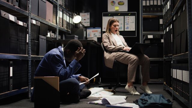 In office space, african american male cop and caucasian female inspector with laptop, examine evidence. Worn-out law officer rests on chair while policeman analyzes statements and clues on ground.