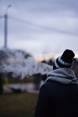 Postać osoby tyłem, w zimowej czapce i szaliku, wydmuchującej chmurę dymu na tle zimowego pochmurnego nieba