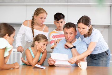 Dad is sitting in kitchen, reading letter and is upset by unpleasant and news. Wife, daughters and children are worried, look carefully at ass and wait for explanation