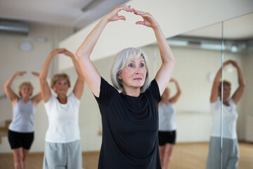 Interested aged woman practicing ballet dance moves during group class in choreographic studio.