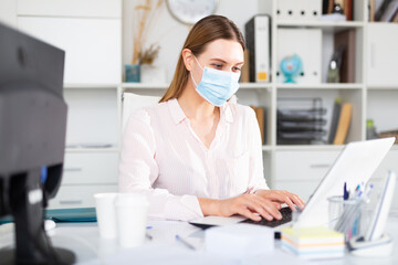 Businesswoman in protective mask working alone with laptop and papers in office, new normal due to...
