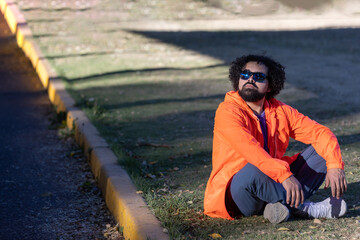Latin man with beard, curly hair, sunglasses and casual clothes, sitting in a park on grass