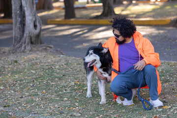 Mexican man with curly hair, beard and wearing casual clothes with sunglasses, sitting in a park hugging his husky dog