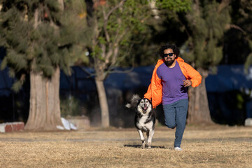 Naklejka na ściany i meble Latin man with beard, curly hair, sunglasses and casual clothes running with his husky dog in a park.