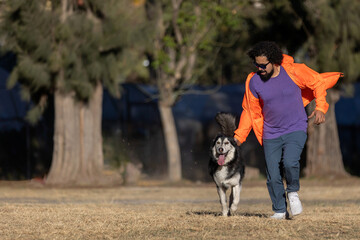 Latin man with beard, curly hair, sunglasses and casual clothes running with his husky dog in a park