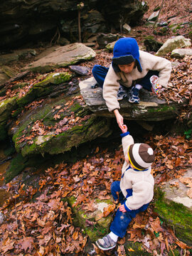 A small boy leans over from atop a rock to hand a snack to his younger brother. Both are wearing tan fleece jackets and winter hats.