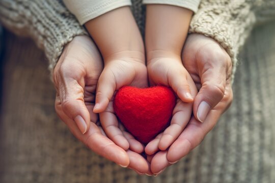 Close-up shot of unrecognizable hands of mother and daughter delicately holding a red heart, photographed from a top-down perspective against a clean and simple backdrop.