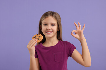 Cute girl with chocolate chip cookie showing OK gesture on purple background