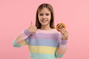 Cute girl with chocolate chip cookie showing thumb up on pink background