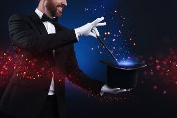 Smiling magician showing trick with wand and top hat on dark blue background, closeup