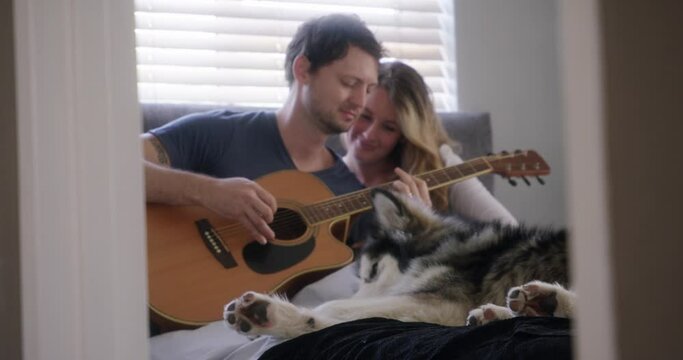 Couple, home and playing guitar with dog for love and bonding, happy in bed with pet. People with domestic animal, husky and musical instrument, time together with trust, commitment and creativity