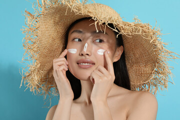 Beautiful young woman in straw hat with sun protection cream on her face against light blue...