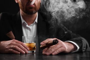 Man with glass of whiskey smoking cigar at dark marble table on black background, closeup - obrazy, fototapety, plakaty