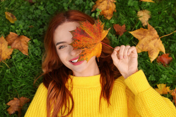 Smiling woman lying on grass and covering eye with autumn leaf, top view