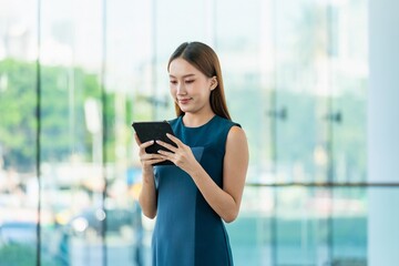 Fototapeta premium Young Asian Woman Smiles Happily While Using Digital Tablet to Manage Her Tasks Remotely While Out Of Office