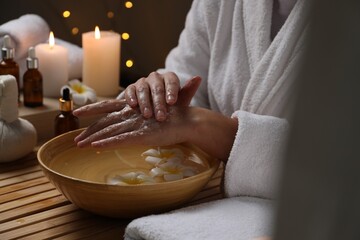Woman applying scrub onto her hands in spa, closeup. Bowl of water and flowers on wooden table