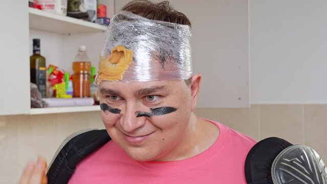 Man with his head wrapped in stretch film, war paint on face, smears peanut butter from jar onto his forehead with finger, challenges him to play fight, attracts Silly fun of father, family leisure
