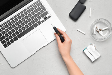 Woman holding modern electronic cigar near laptop on grey background