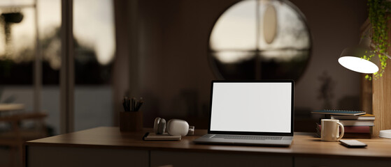 A cosy, modern home workspace at night with a white-screen laptop computer mockup on a wooden desk.
