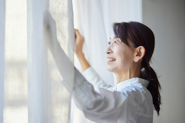 リビングのカーテンを開ける日本人のシニア女性 - obrazy, fototapety, plakaty