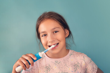 Beauty portrait of happy cheerful positive beautiful girl, teen is brushing her healthy white teeth with electric toothbrush with toothpaste, looking at camera and smiling on blue background.