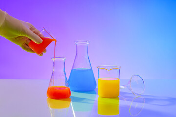 A hand is holding a beaker pouring red liquid from it through an erlenmeyer flask, next to it are two experimental glass flasks containing yellow and blue liquids on a gradient background.