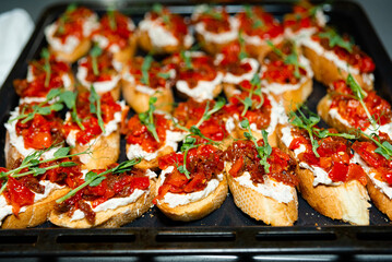 Bruschetta with Tomato, Cheese, and Arugula on Tray. Toasted bruschetta topped with creamy cheese, sun-dried tomatoes, and fresh arugula arranged neatly on a black baking tray.