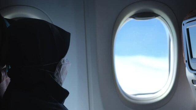 A veiled woman wearing a mask looking out the window from the seat of an airplane in flight 