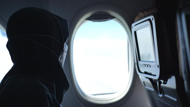 A veiled woman wearing a mask looks out the window from the seat of an airplane that is flying and shaking