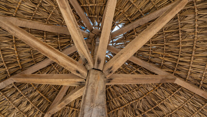 A fragment of a solar umbrella. Close-up. Bottom view. Full frame. Straw is visible intertwined in a circle, wooden reinforcing crossbars. The blue sky shines through the holes. Madagascar