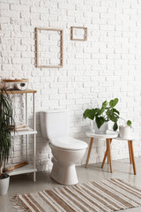 Interior of light restroom with ceramic toilet bowl, shelving unit and houseplant near white brick...
