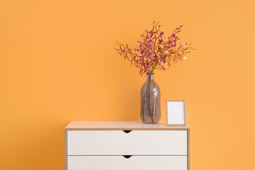 Vase with beautiful flowers and blank frame on chest of drawers near orange wall