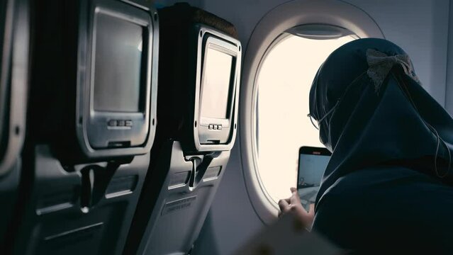 Asian woman in hijab looking at the shaking airplane window while recording with her phone during landing