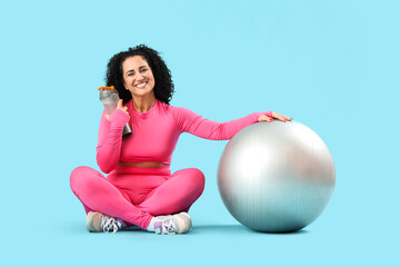 Sporty adult woman with fitball and bottle of water on blue background