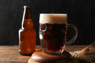 Mug and bottle of cold dark beer with wheat on wooden table against black grunge background