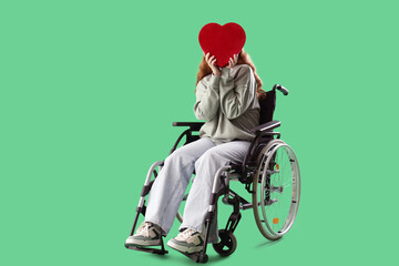 Young redhead woman in wheelchair with heart-shaped gift box on green background
