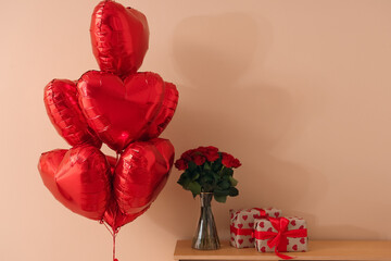 Beautiful bouquet of roses with heart-shaped balloons and gift boxes on commode near beige wall....
