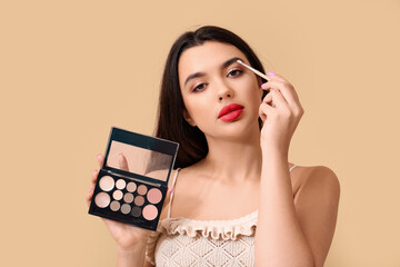 Beautiful young woman with makeup brush and stylish eyeshadow palettes on beige background - obrazy, fototapety, plakaty
