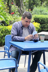 Fototapeta premium 40-year-old dark-skinned Latino man drinks a coffee and uses his cell phone on a park bench to work remotely, make calls, send messages, buy and pay online