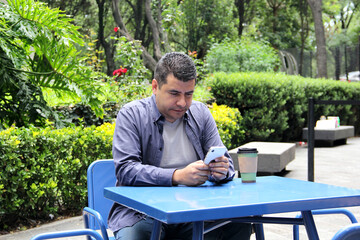Fototapeta premium 40-year-old dark-skinned Latino man drinks a coffee and uses his cell phone on a park bench to work remotely, make calls, send messages, buy and pay online