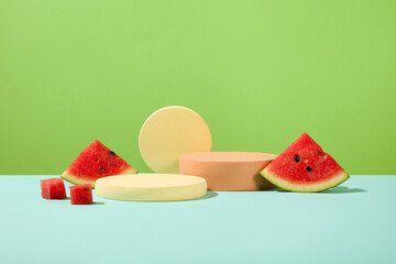 Watermelon slices are placed next to cylindrical platforms on a blue surface with a green background. Ideal space for displaying products with main ingredients from watermelon.