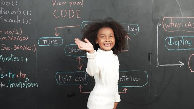 African girl smiling and waving at camera while standing at board with engineering prompt or coding, programing system written in STEM classroom. Child looking at camera while greeting. Erudition.