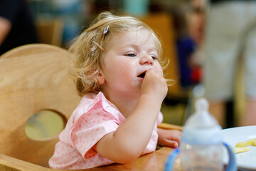 Fototapeta premium Adorable toddler girl eating healthy vegetables and unhealthy french fries potatoes. Cute happy baby child taking food from dish at daycare or nursery canteen.
