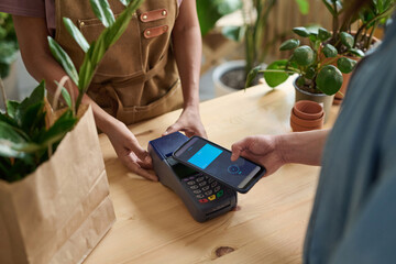 Customer paying for her purchase in plant store with smartphone, cashier receiving payment