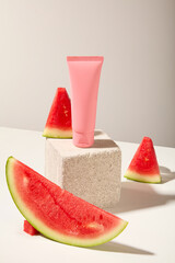On a white background, a pink plastic tube displayed on stone podium with fresh juicy watermelon slices. Front view, mockup scene for advertising cosmetic of watermelon ingredient
