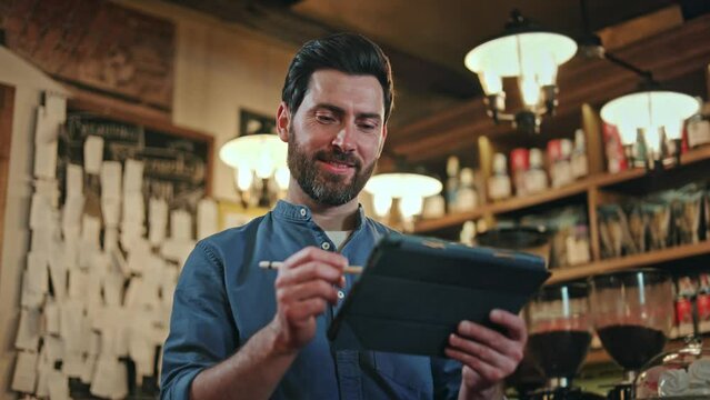 Friendly caucasian man in blue uniform adding information into digital tablet during working process at bar. Competent staff member with stylus pen in hand taking order from guest at modern workplace.